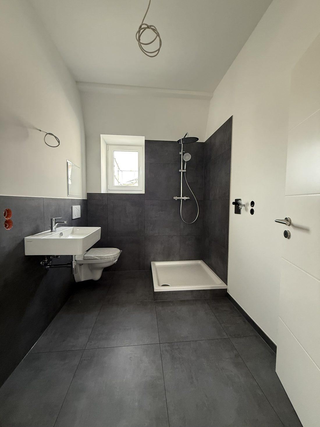 Tageslichtbad mit Fenster, dunklen Wandfliesen, bodengleicher Dusche, Hänge-WC und Waschtisch