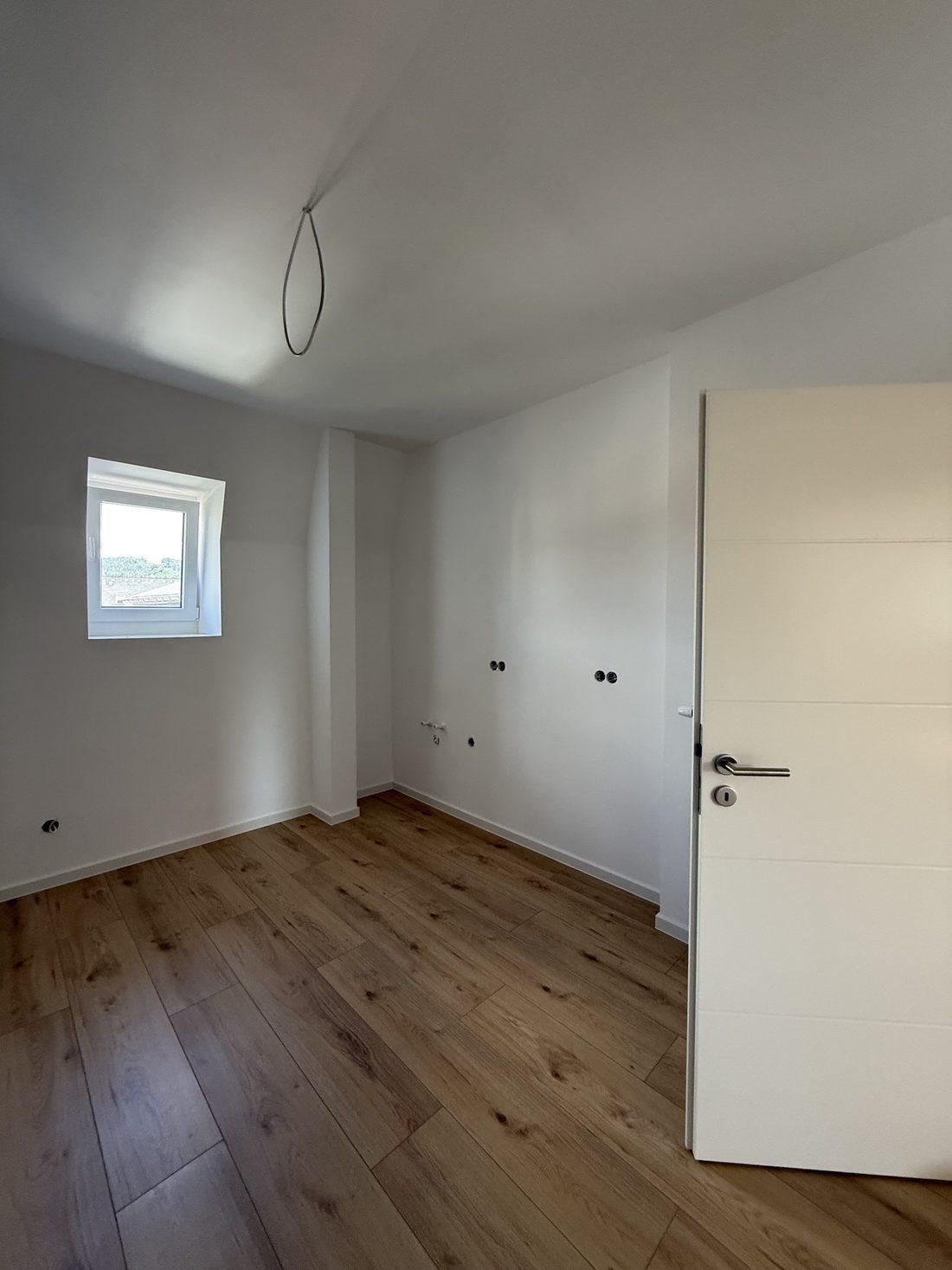 Küche mit Fenster, offen stehender Zimmertür, Anschlüssen an der Wand und Boden in Eichenoptik